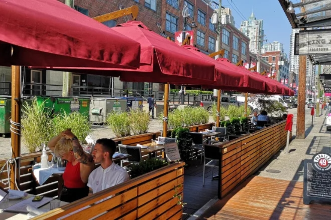 people seated in a curbside restaurant patio covered by red umbrellas above